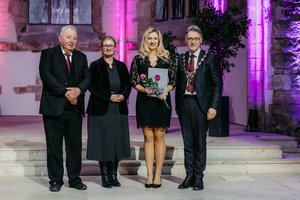 Four people are standing side by side on a stage in a festively lit church. The person in the middle is holding a certificate and a bouquet of flowers. The stone walls and purple lighting in the background can be seen.