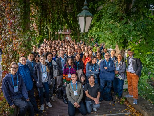 A large group of around 200 participants at the Learning & Memory Conference 2025 stand close together in the countryside under a pergola, smiling at the camera. Researchers of various nationalities can be seen in the foreground and background.