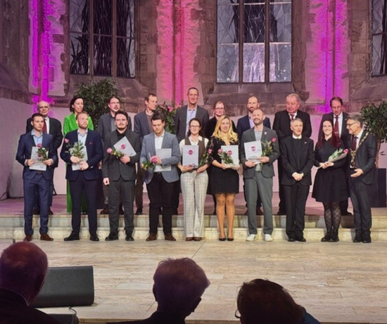 Group photo of the doctoral award winners from Otto von Guericke University Magdeburg at the ceremony in St Johannes Church. A large group stands on a stage in front of illuminated church windows; they are all holding certificates and flowers.