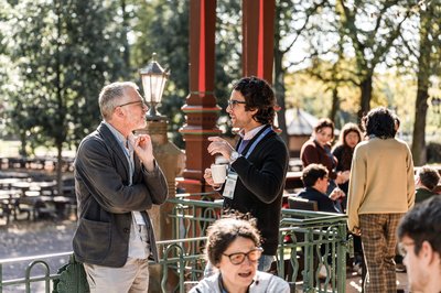 Several conference participants chat in the sunshine over coffee on a terrace with green metal railings. Two men talking stand in the foreground, with other groups in the background in a relaxed atmosphere.