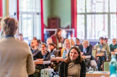 Conference participants sit in a bright, wood-paneled hall and listen attentively to a speaker. In the foreground, a woman in the audience is seen smiling and focused.