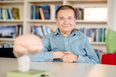 [Translate to English:] Ein Mann mittleren Alters im karierten Hemd sitzt an einem Tisch in einem Bücherzimmer und lächelt in die Kamera. Im Vordergrund unscharf ein Modell eines Gehirns.