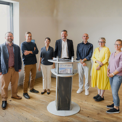 [Translate to English:] Sieben Personen stehen bei einem Treffen in einem hellen Raum um einen Stehtisch mit LIN-Aufsteller.